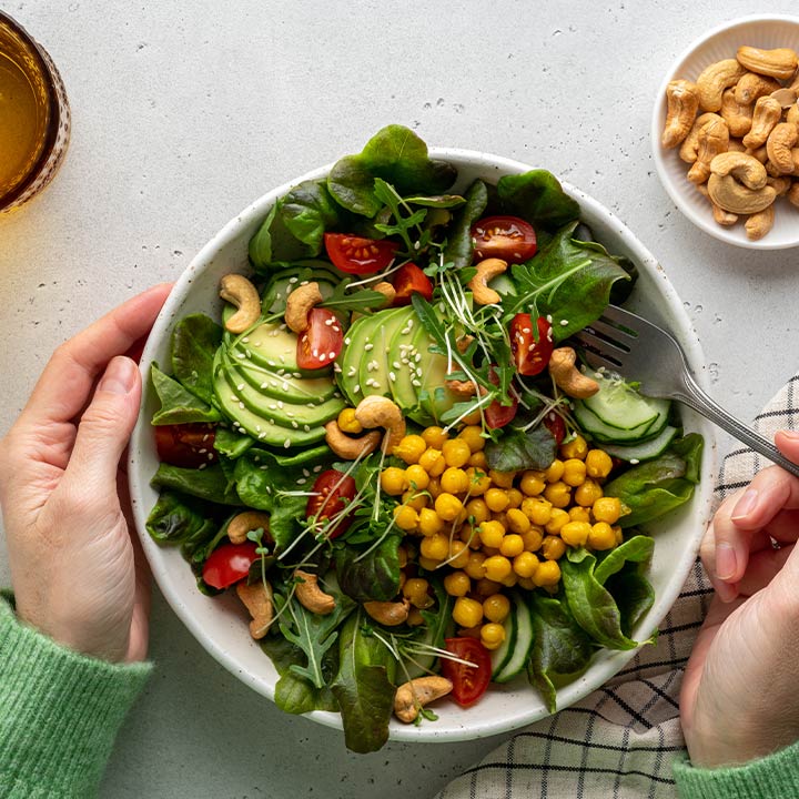 Anonymous female dining healthy vegan plant-based salad in bowl with avocado, cashew, micro-greens, pok choi, chickpeas, tomato, lettuce, cucumber, sesame. Flexitarian fatty acids and dietary fiber.